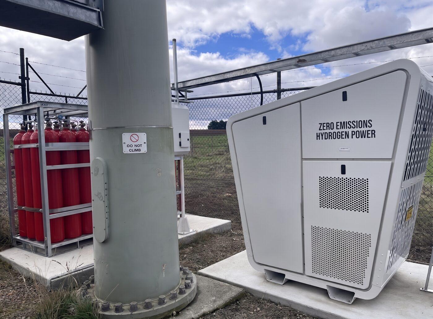 Een witte waterstofgenerator met het opschrift Zero Emissions Hydrogen Power, opgesteld naast een rek gascilinders.